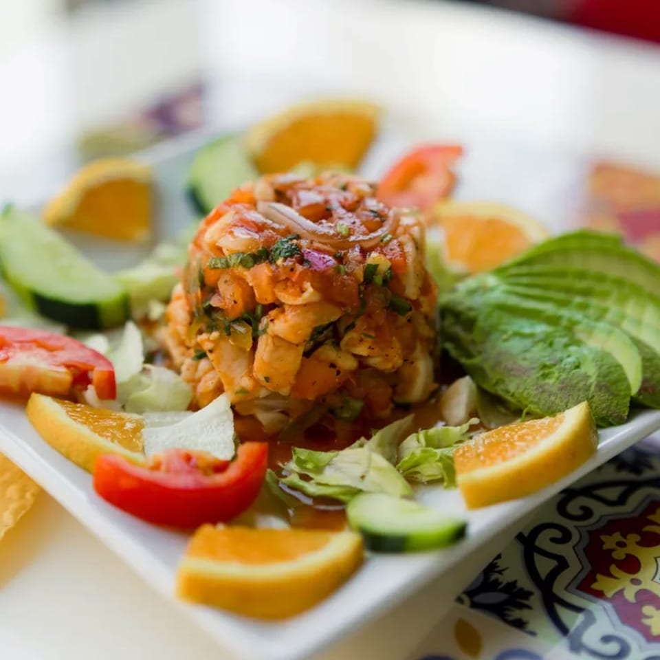 A vibrant ceviche dish with shrimp, avocado, oranges, tomatoes, and cucumbers on a white plate.