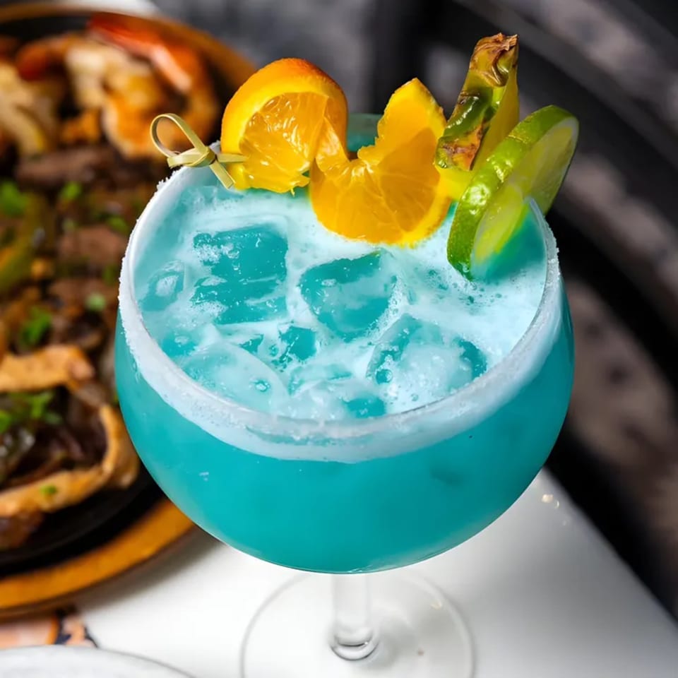 A vibrant blue margarita with ice, garnished with orange, pineapple, and lime slices.