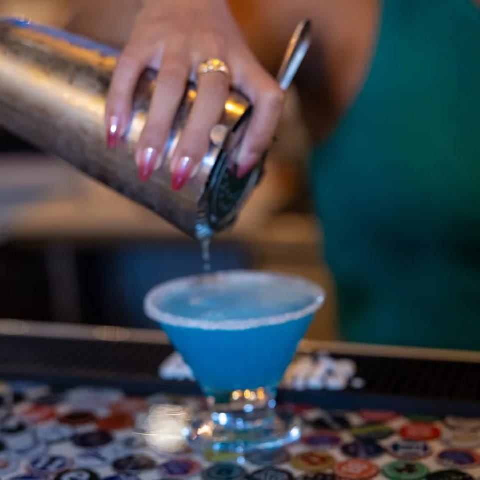 A bartender pours a vibrant blue margarita from a shaker into a salt-rimmed glass.