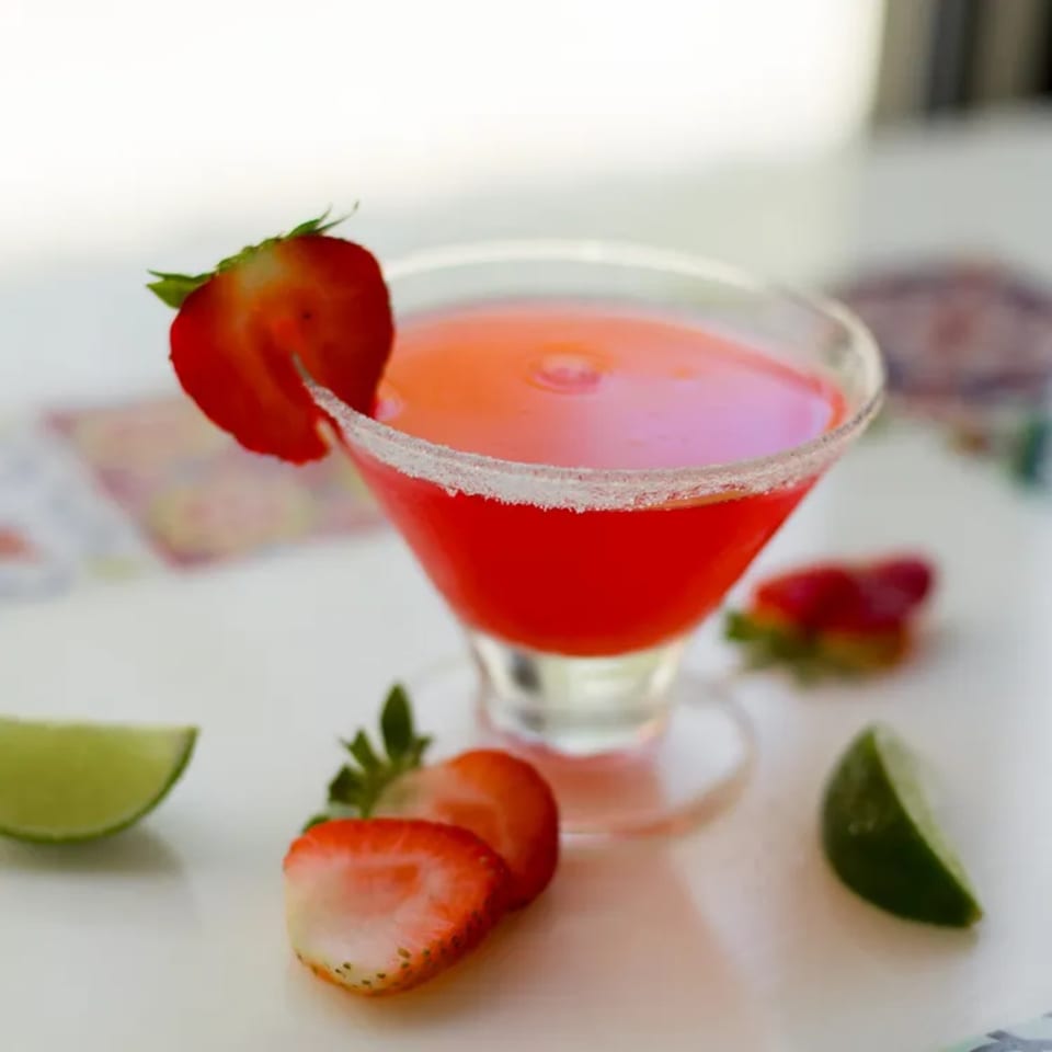 A vibrant red strawberry margarita in a sugar-rimmed glass, garnished with fresh strawberries and lime wedges.