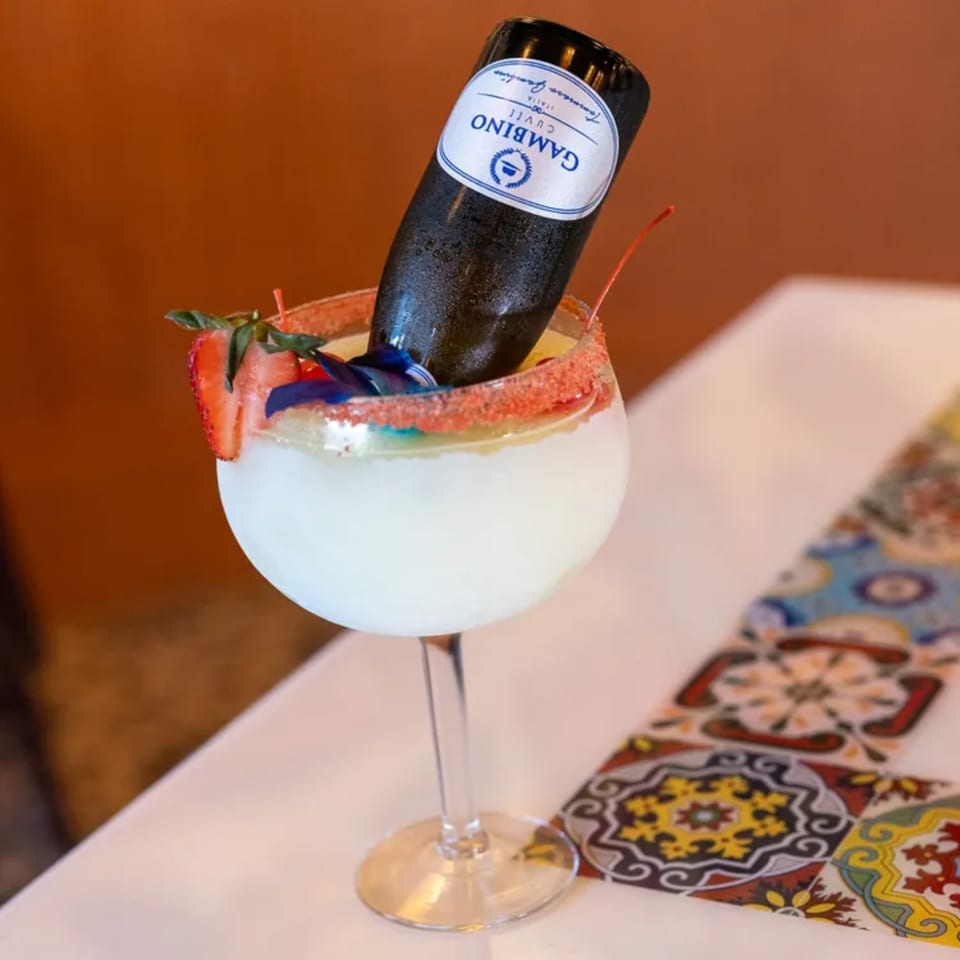 A large margarita glass with a mini bottle of Gambino Prosecco, strawberry, and blue garnish.